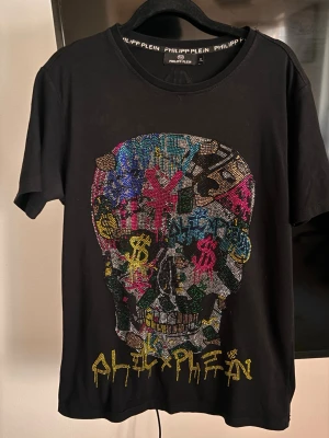 Philipp Plein t-shirt - Svart t-shirt från Philipp Plein med färgglad döskalle framtill dekorerad med glittriga stenar och logga i gult nedtill. Rund halsringning och kort ärm. Materialet är mjuk bomull och passformen är normal.