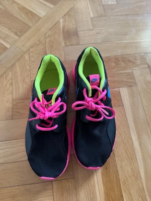 Nike sneakers svart/blå/rosa - Nike sneakers med svart mesh och syntetmaterial, blå swoosh på sidan, neonrosa snören och sula samt limegrön insida. Snygg och sportig modell med lågt skaft, perfekt för dig som gillar färgstarka detaljer.