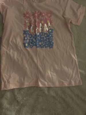 Rosa t-shirt med blommigt tryck - Söt rosa t-shirt med korta ärmar och blommigt tryck i rosa och blått framtill. Texten 'DREAM' står i vitt över blommorna. Perfekt för dig som gillar färg och mönster.