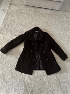 Svart dubbelknäppt kappa Esprit - Stilren svart kappa från Esprit med dubbelknäppning och bred krage. Kappan har två fickor framtill och är helfodrad. Klassisk modell som passar perfekt till höst och vinter. Tillverkad i ett ullblandat material för extra värme.