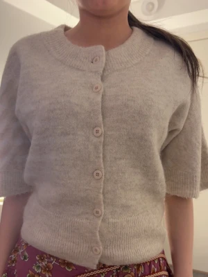 Beige kofta med kort ärm och knappar - Mysig beige kofta med korta ärmar och rund halsringning. Koftan har fem knappar framtill och är stickad i ett mjukt material. Perfekt för lager-på-lager och enkel att matcha med olika outfits.