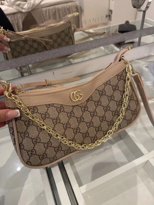  beige väska  - Snygg handväska från  i beige canvas med klassiskt GG-mönster och ljusbruna läderdetaljer. Väskan har en guldfärgad kedja som axelrem och GG-logo framtill. Perfekt accessoar för att lyfta din outfit.