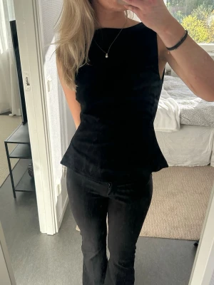 Svart ärmlös topp med utsvängd midja - Snygg svart ärmlös topp med hög halsringning och lätt utsvängd midja. Toppen har en stilren och enkel design som passar perfekt till både jeans och kjol. Materialet ser ut att vara mjukt och följsamt, vilket ger en bekväm passform.