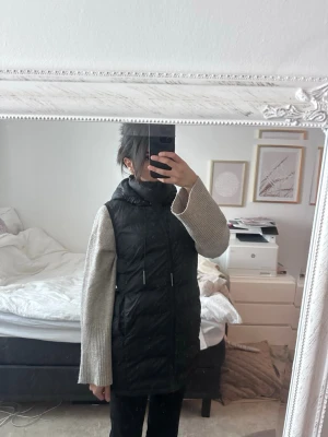 Svart lång dunväst med huva - Snygg svart lång dunväst med dragsko i midjan och huva. Västen har en stilren look och är perfekt att slänga över en stickad tröja. Passar dig som gillar en avslappnad men trendig stil.