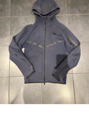 Mörkblå Nike Tech Fleece hoodie - Nike Tech Fleece hoodie i mörkblått med hel dragkedja, huva och diskret Nike-logga på bröstet. Har en snygg detalj med diagonal söm och en ficka med dragkedja på ärmen. Tillverkad i mjukt och bekvämt fleecematerial.