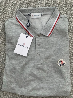 Grå pikétröja från Moncler - Snygg grå pikétröja från Moncler med klassisk krage och tre knappar framtill. Kragen har vita och röda ränder för en sportig touch och bröstet pryds av Monclers ikoniska logotyp. Perfekt till jeans eller shorts för en clean look.