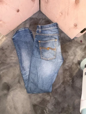 Blå jeans från Nudie Jeans - Säljer ett par klassiska blå jeans från Nudie Jeans med snygga slitningar och kontrastsömmar. Jeansen har fem fickor och en broderad detalj på bakfickan. Perfekta för en avslappnad och trendig look.