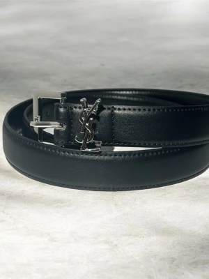 Svart skärp från Saint Laurent - Stilrent svart skärp från Saint Laurent i skinn med klassiskt YSL-logo i metall framtill. Skärpet har en silverfärgad spänne och diskreta sömmar längs kanterna. Perfekt accessoar för att lyfta din outfit.