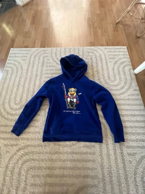 Blå Polo Bear hoodie från Ralph Lauren - Blå hoodie från Ralph Lauren med Polo Bear-tryck framtill. Hoodien har huva och lång ärm, samt ett färgglatt motiv av en björn med skidutrustning. Mjuk bomullskänsla och klassisk passform. Perfekt för en avslappnad och cool stil.