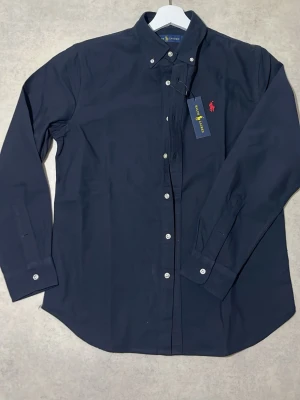 Ralph Lauren - Stilren och klassisk skjorta från Ralph Lauren i mörkblå färg med ikonisk röd broderad logga på bröstet. Perfekt både till vardag och lite mer uppklätt.