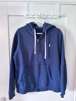 Marinblå hoodie från Polo Ralph Lauren - Snygg marinblå hoodie med dragkedja från Polo Ralph Lauren. Klassisk design med vit logga på bröstet, vita dragsnören och två stora fickor framtill. Tillverkad i mjuk bomullsmix och har huva för extra komfort.