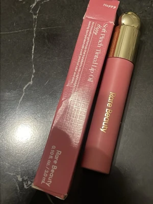 Rare Beauty Soft Pinch Tinted Lip Oil - Rare Beauty Soft Pinch Tinted Lip Oil i nyansen Happy. Läppoljan har en mjuk rosa färg och kommer i en stilren tub med guldfärgat lock. Ger en lätt tint och glans till läpparna. Förpackningen rymmer 3 ml.