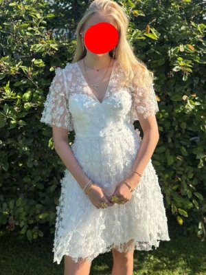 Vit spetsklänning med v-ringning - Superfin vit klänning med v-ringning och korta ärmar i spets. Klänningen har blommiga spetsdetaljer. 