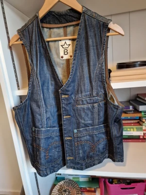 Jeansväst från B Brand med fickor - Snygg jeansväst från B Brand i klassisk blå denim. Västen har flera stora fickor framtill, dekorativa sömmar och en cool, avslappnad look. Perfekt att slänga över en t-shirt för en extra edge. Märke med krona och B på insidan.