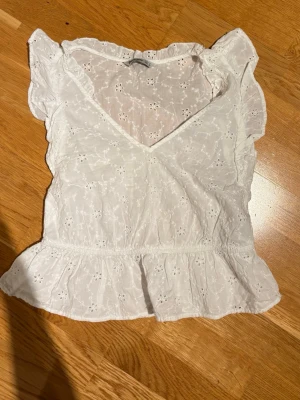 Vit blus med broderade blommor - Söt vit blus från Stradivarius med broderade blommor och volangdetaljer vid ärmar och nederkant. Blusen har v-ringning och är tillverkad i bomull, perfekt för en fräsch och somrig look.