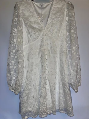 Vit blom broderad klänning - Superfin vit klänning ifrån Bubbleroom med broderade blommor och transparenta långärmade ärmar. Klänningen har v-ringning, knappar framtill. Perfekt för sommarens alla tillfällen! Helt ny med sina prislappar kvar.