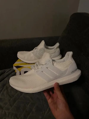 Adidas Ultra Boost vita sneakers - Fräscha helvita Adidas Ultra Boost sneakers med stickad ovandel och klassiska tre ränder på sidan. Skorna har en chunky Boost-sula för maxad komfort och sportig vibe. Perfekta för dig som gillar stilrena och cleana sneakers med modern känsla.