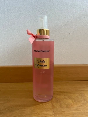 Women'secret Daily Romance Body Mist - Fräsch body mist från Women'secret i doften Daily Romance. Flaskan är rosa med gulddetaljer och en söt rosa rosett vid halsen. Praktisk sprayflaska i plast, perfekt att ha med i väskan för en snabb uppfräschning.