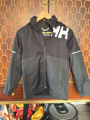 Svart vindjacka från Helly Hansen - Svart vindjacka från Helly Hansen med vit HH-logga på axeln, hel dragkedja och huva. Jackan har två fickor framtill, reflekterande detaljer på ärmarna och meshfoder på insidan. Perfekt för blåsiga dagar och enkel att ta med sig.