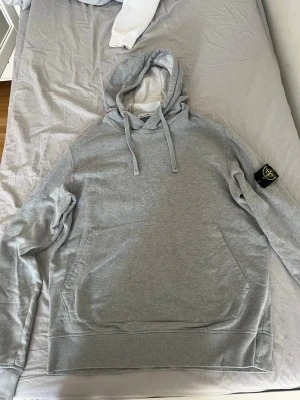 Grå hoodie från Stone Island - Snygg grå hoodie från Stone Island med klassisk logga på vänster ärm. Hoodien har huva med dragsko och en stor magficka framtill. Perfekt för en avslappnad och trendig stil. Mjuk bomullskänsla och stilren design.  Storlek L blev liten på mig 