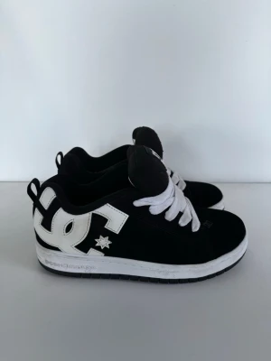 Svarta DC sneakers med vit logga - Svarta sneakers från DC Shoes med stor vit DC-logga på sidan och vita skosnören. Skorna har en klassisk låg profil och tjock vit sula. Ovandelen är i mockaliknande material och tungan har DC-logga i vitt. Perfekta för en streetwear-look.