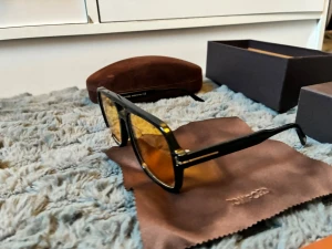 Svarta Tom Ford solglasögon - Snygga solglasögon från Tom Ford med svart, kantig båge och gulbruna glas. Klassisk design med diskreta gulddetaljer vid gångjärnen. Kommer med originalfodral och putsduk. Perfekta för dig som vill ha en stilren accessoar. Den är i nytt skick men har ett litet jack på hög sida inne från mer en så är den ny 