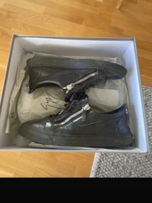Svarta sneakers från Giuseppe Zanotti - Snygga svarta sneakers i läder från Giuseppe Zanotti med coola silverfärgade dragkedjor på sidorna. Skorna har lågt skaft och klassisk snörning. Perfekta för dig som gillar en stilren men edgy look. De ända är hälen som ni ser på bilderna som är ganska andvända annars är skorna bara smutsiga, denna modell är från 2014 så de kommer ingen logga på toeboxen kvitto kommer med!
