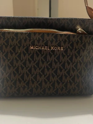  brun monogram handväska - Snygg handväska i brunt konstläder med klassiskt MK-monogram över hela väskan. Väskan har guldfärgade detaljer, dragkedja och ett handtag i ljusbrunt läder. Perfekt för dig som gillar stilrena accessoarer med lyxig känsla.