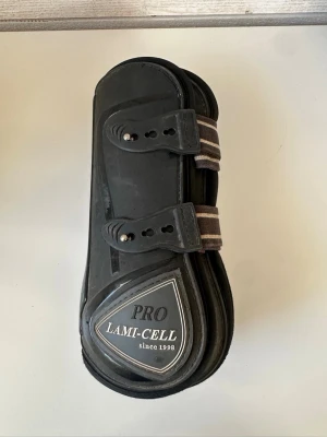 Svarta benskydd Lami-Cell - Svarta benskydd från Lami-Cell Pro med två elastiska remmar i brunt och vitt. Tillverkade i slitstarkt plastmaterial och vadderade på insidan. Perfekta för hästsport och skydd av hästens ben vid ridning eller träning. Sparsamt använda men säljer då dem inte passade.