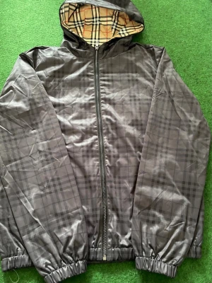 Windbreaker burberry - Snygg grå vindjacka med diskret rutmönster och dragkedja framtill. Jackan har huva med klassiskt beige-rutigt foder och elastiska muddar vid ärmslut och nederkant. Perfekt för blåsiga dagar och enkel att matcha med olika outfits.
