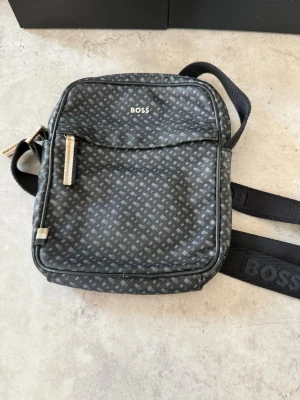 Svart axelväska från BOSS - Snygg svart axelväska från BOSS med diskret mönster i grått och justerbar axelrem. Väskan har dragkedja och en praktisk ytterficka framtill. Tillverkad i slitstarkt material med detaljer i metall och logga på både väska och rem.