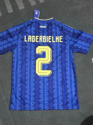 Blå Sverige fotbollströja Adidas - Säljer en blå fotbollströja från Adidas med Sveriges landslagsemblem och gula detaljer. Tröjan har trycket 'Lagerbielke' och nummer 2 på ryggen, samt gula ränder på axlarna. Tillverkad i polyester med retroinspirerat mönster och korta ärmar.