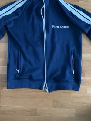Blå zip-jacka från Palm Angels - Snygg blå zip-jacka från Palm Angels med vita ränder längs ärmarna och vit dragkedja. Jackan har två fickor med dragkedja och logga på bröstet. Perfekt för en sportig och trendig look.