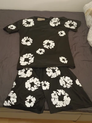 Svart set från Denim Tears med blommor - Säljer ett svart set från Denim Tears med t-shirt och shorts. Setet har stora vita blommönster tryckta över hela plaggen. Materialet är mjukt och följsamt, perfekt för en avslappnad stil. Märket syns tydligt i nacken på tröjan.