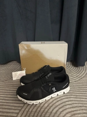 Svarta On Running sneakers med snörning - Snygga svarta sneakers från On Running med vit, unik sula och diskret logga på sidan. Skorna har snörning och är tillverkade i ett lätt syntetmaterial som andas. Perfekta för dig som gillar sportig och modern stil.