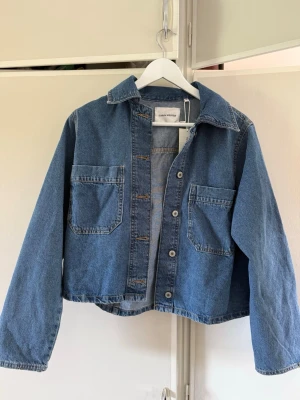 Helt ny jeansjacka från Carin Wester - Snygg blå jeansjacka från Carin Wester i klassisk modell med två stora bröstfickor och knappar framtill. Jackan har en rak passform och krage, perfekt för en avslappnad look. Tillverkad i slitstarkt jeansmaterial.