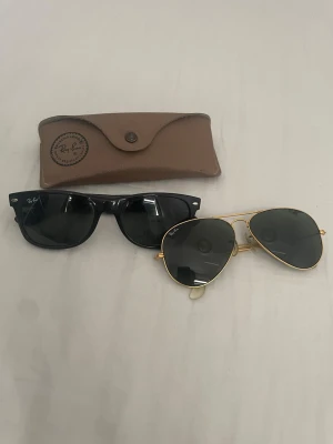 Ray-Ban solglasögon Wayfarer & Aviator - Två par klassiska Ray-Ban solglasögon: ett par svarta Wayfarer med plastbågar och ett par Aviator med tunna guldfärgade metallbågar och mörka linser. Medföljer gör ett brunt fodral med Ray-Ban-logga. Perfekt för dig som vill ha ikoniska modeller.