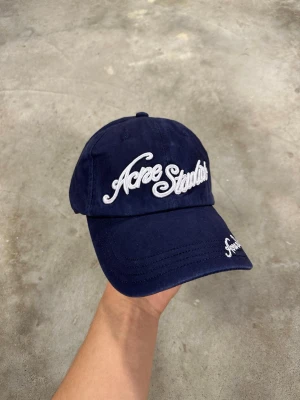 Mörkblå keps från Acne Studios - Snygg mörkblå keps från Acne Studios med vit broderad logga framtill och på skärmen. Klassisk böjd skärm och justerbar passform bak. Perfekt accessoar för en cool och avslappnad stil.