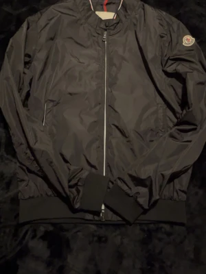 Moncler Reppe jacka - Svart jacka från Moncler med klassisk rund halsringning och ribbade muddar. Jackan har dragkedja framtill, två sidofickor och Moncler-logga på vänster ärm. Materialet är glansigt och tunt, perfekt för lager-på-lager. Enkel och stilren design som funkar året runt.