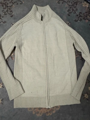 Beige stickad ziptröja med hög krage - Beige stickad tröja med dragkedja framtill och hög krage. Tröjan har ribbstickade partier längs ärmar, sidor och nederkant för en snygg och stilren look. Perfekt för lager-på-lager och passar till många outfits.