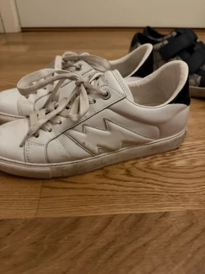 Vita Zadig sneakers med blixt detalj  - Snygga vita sneakers från Zadig & Voltaire med ikonisk blixtdetalj på sidan och svart hälparti. Skorna har klassisk låg modell, vita snören och är tillverkade i läder. Perfekta för en stilren och trendig look.