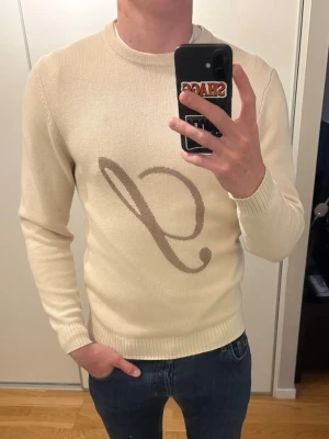 Beige stickad tröja från Stenströms - Snygg beige stickad tröja med en stor brun logga  på bröstet. Tröjan har rund halsringning, långa ärmar och ribbade muddar. Perfekt för dig som gillar stilrena och trendiga plagg med en twist.