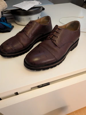 Bruna loafers i läder med grov sula - Säljer ett par bruna loafers i äkta läder med snyggt texturerad yta och markerad tåkappa. Skorna har en rejäl, svart gummisula med grovt mönster för extra grepp. Klassisk design med snörning och låg profil, perfekta för en stilren look.