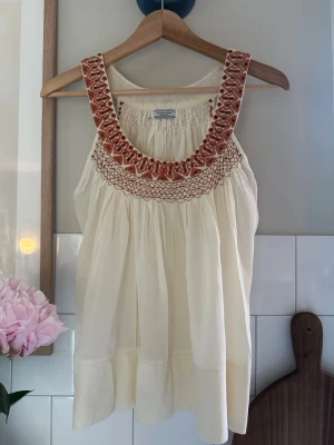 Broderad topp från Hunkydory - Superfin ärmlös linnetopp från Hunkydory i ljusbeige med breda axelband och broderade detaljer i rostbrunt och vitt runt ringningen. Toppen har en luftig passform och smockad rygg för extra komfort. Lite oklar stlk men passar en m/l beroende på passform. Sparsamt använd. 