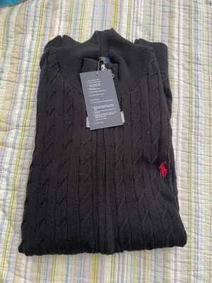 Svart kabelstickad ziptröja Polo Ralph Lauren - Svart kabelstickad ziptröja från Polo Ralph Lauren med röd broderad logga på bröstet. Tröjan har hög krage och dragkedja framtill. Klassisk och stilren design, perfekt för lager-på-lager. Materialet är mjukt och bekvämt.
