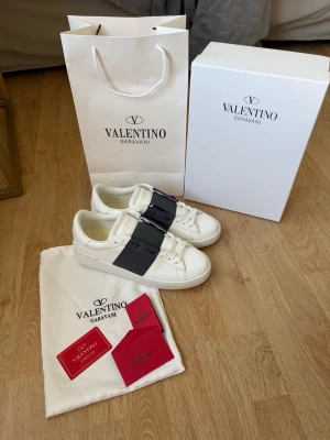 Valentino Garavani Opens vita sneakers - Stilrena vita sneakers från Valentino Garavani med bred mörkblå rem över vristen och klassisk logga på plösen. Skorna har en chunky vit sula med rutmönster undertill och är tillverkade i ett fint material. Perfekta för dig som vill ha en lyxig och trendig look nu i sommar. De är något små i storleken. De är helt nya och aldrig använda, skriv vid frågor eller funderingar. 