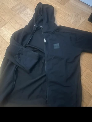 Svart hoodie med dragkedja från ICANIWILL - Snygg svart hoodie från ICANIWILL med dragkedja och huva. Mjuk bomullskänsla och stilren design med logga på bröstet. Perfekt för chill eller träning. Klassisk passform och ribbade muddar vid ärmslut.