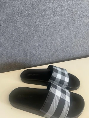 Burberry rutiga svarta tofflor - Snygga svarta tofflor från Burberry med klassiskt rutigt mönster i svart, vitt och grått över remmen. Tofflorna har en enkel slip-on design och är tillverkade i syntetmaterial med en bekväm fotbädd. Perfekta för chill dagar hemma eller till stranden.
