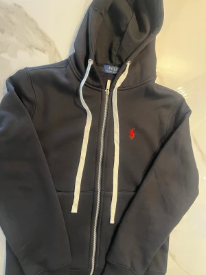 Svart hoodie från Polo Ralph Lauren - Säljer en svart hoodie från Polo Ralph Lauren med dragkedja, vit dragsko och den klassiska röda loggan broderad på bröstet. Hoodien har fickor framtill och är fodrad för extra komfort. Perfekt för dig som vill ha en stilren och bekväm look.