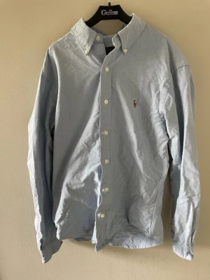 Ljusblå skjorta från Ralph Lauren - Klassisk ljusblå skjorta från Ralph Lauren med button-down krage och broderad logga på bröstet. Skjortan har långa ärmar och knäppning framtill. Perfekt för en stilren och avslappnad look.